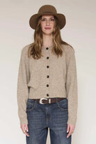 A woman wearing a beige button-front cardigan, blue jeans, a brown belt, and a brown wide-brim hat, standing against a plain light background.