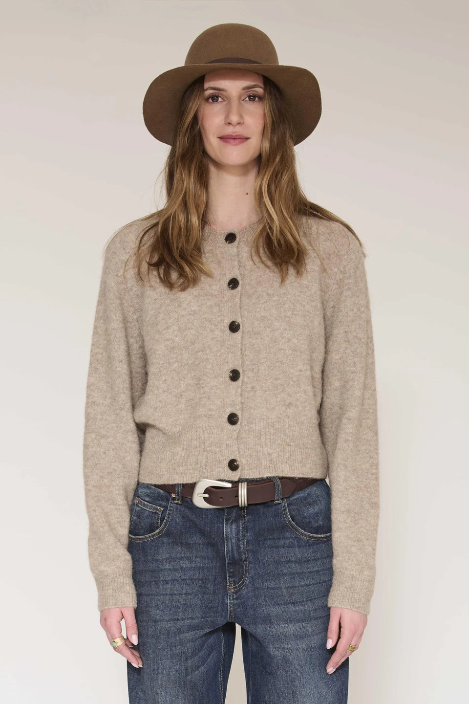 A woman wearing a beige button-front cardigan, blue jeans, a brown belt, and a brown wide-brim hat, standing against a plain light background.