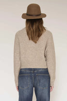 A woman shown from the back wearing a beige knit sweater, blue jeans, and a brown felt hat against a plain light background.