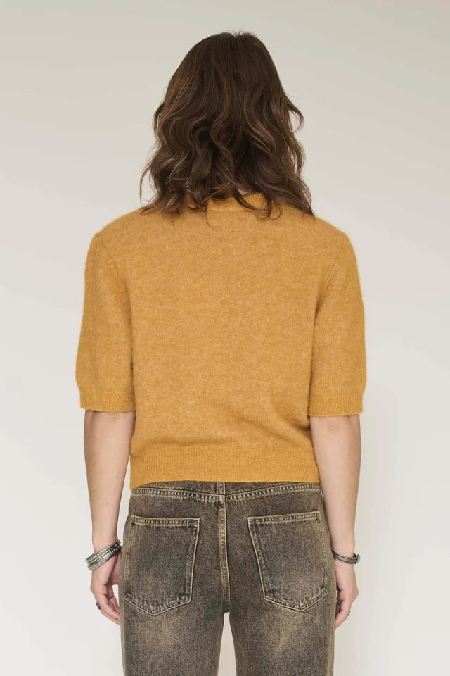 A woman viewed from the back wearing a mustard yellow short-sleeve knit top and dark gray jeans against a plain light background.
