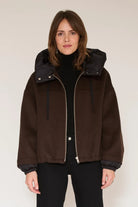 A woman wearing a dark brown suede-like hooded jacket with a black inner hood and black turtleneck, standing against a plain light beige studio background.