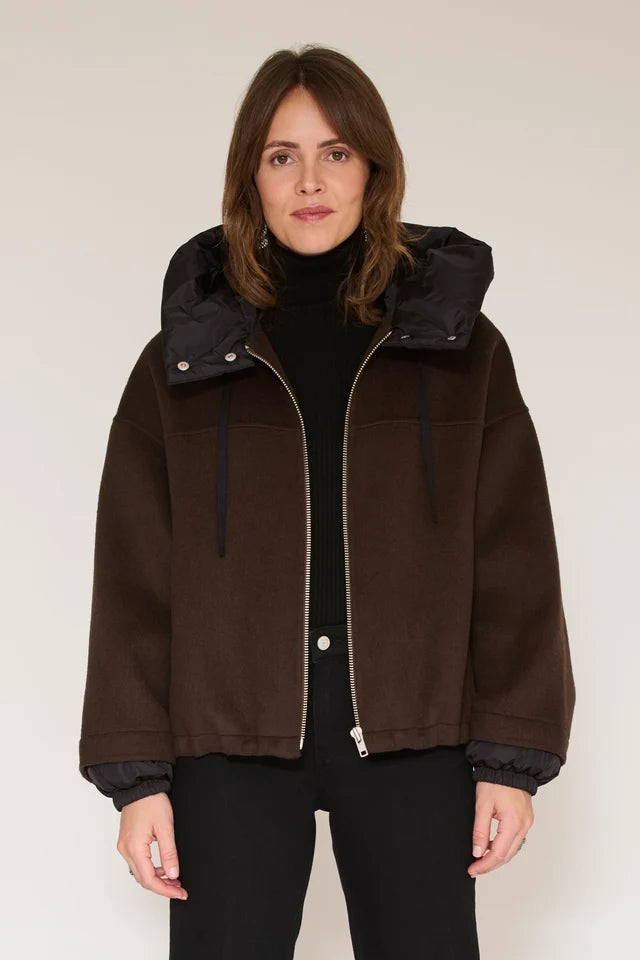 A woman wearing a dark brown suede-like hooded jacket with a black inner hood and black turtleneck, standing against a plain light beige studio background.