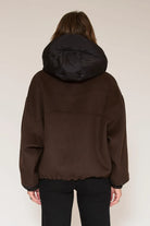 A person viewed from the back wearing a dark brown hooded jacket with a black hood and black pants, standing against a plain light beige studio background.