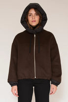 A woman wearing a dark brown zip-up hooded jacket with a black inner hood lining and black cuffs, paired with black pants, against a plain light beige studio background.