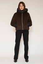 A model stands against a plain light background wearing a dark brown hooded jacket with a black front zipper and black gloves, paired with black pants and black shoes.