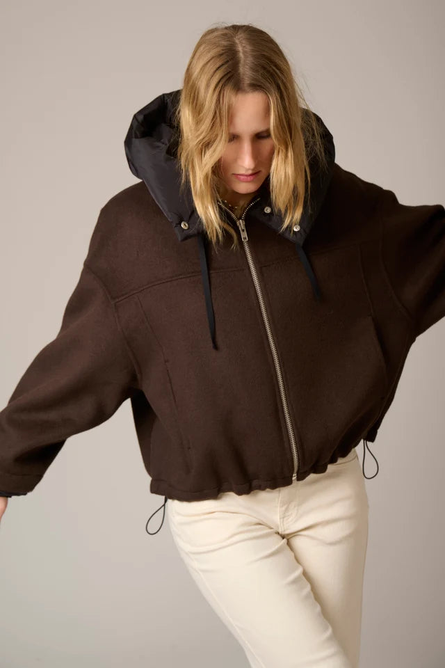 A woman wearing a dark brown cropped hooded jacket with a black inner hood and white pants, posed against a plain light gray studio background.