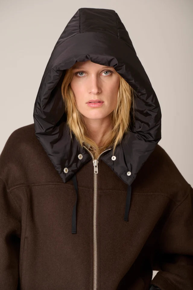 A model wearing a dark brown zip-up hoodie with a large black puffer hood attached, photographed against a plain light beige studio background.
