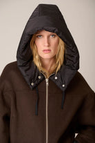 A model wearing a dark brown zip-up hoodie with a large black puffer hood attached, photographed against a plain light beige studio background.