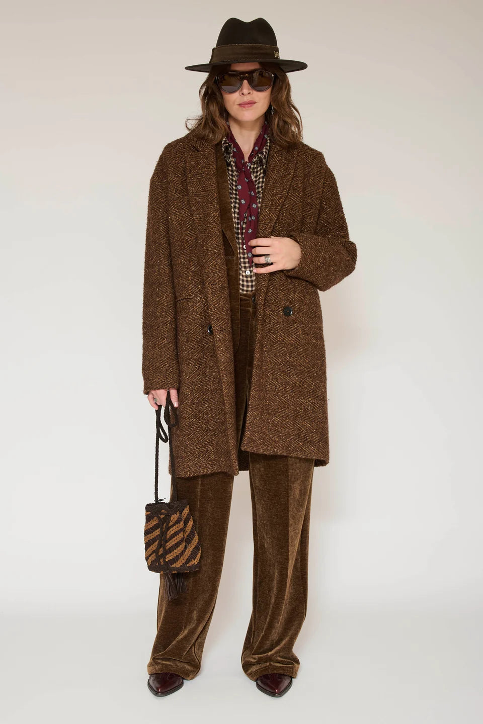 A model wearing a brown tweed double-breasted coat, matching brown velvet wide-leg trousers, a patterned scarf, a dark brown fedora hat, and sunglasses, holding a small brown and black striped handbag against a plain light background.
