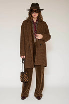 A model wearing a brown tweed double-breasted coat, matching brown velvet wide-leg trousers, a patterned scarf, a dark brown fedora hat, and sunglasses, holding a small brown and black striped handbag against a plain light background.