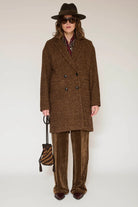 A model wearing a brown double-breasted tweed coat, matching brown velvet trousers, a brown fedora hat, sunglasses, and holding a brown and black striped bucket bag against a plain light background.