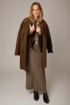 A woman modeling a brown textured coat over a brown and beige checkered skirt, with black boots, against a neutral studio background.