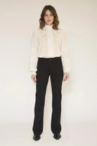 A woman stands against a plain light background wearing a cream long-sleeve blouse with ornate embroidery around the high neck and cuffs, paired with black pants and black shoes.