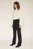 A model wearing a white long-sleeve blouse with gold embroidery and black trousers, standing against a plain off-white studio background.