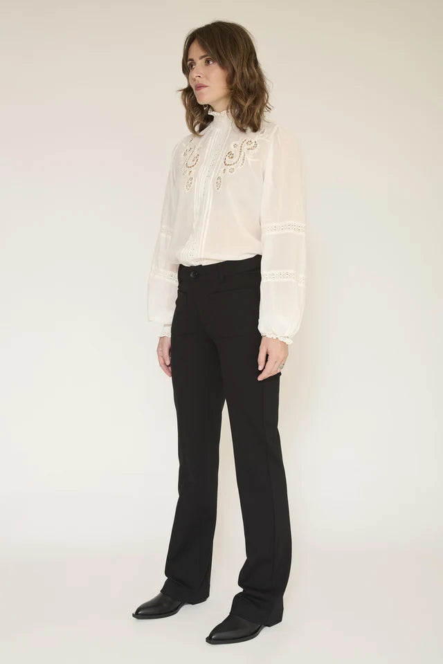 A model wearing a white long-sleeve blouse with gold embroidery and black trousers, standing against a plain off-white studio background.