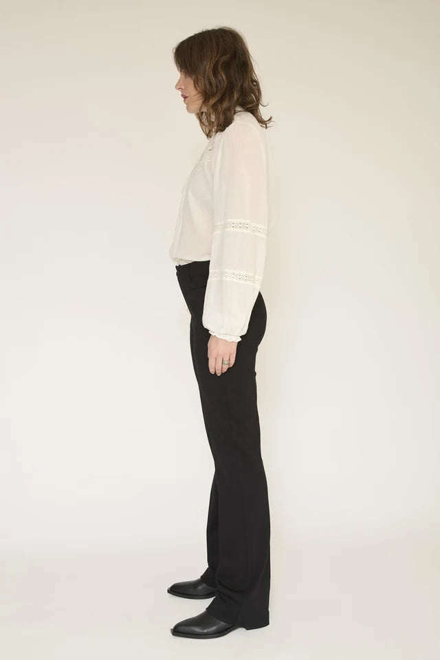 A woman shown in profile wearing a white long-sleeve blouse with lace detailing on the cuffs and black wide-leg trousers, standing against a plain off-white studio background.