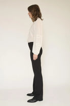 A woman shown in profile wearing a white long-sleeve blouse with lace detailing on the cuffs and black wide-leg trousers, standing against a plain off-white studio background.