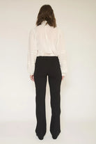 A woman shown from the back wearing a white long-sleeve blouse with sheer sleeves and black high-waisted flared trousers, photographed against a plain light background.