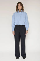 A model stands against a plain light background wearing a light blue long-sleeve button-up shirt and dark navy pinstriped trousers.
