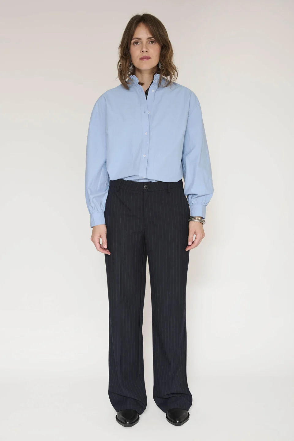 A model stands against a plain light background wearing a light blue long-sleeve button-up shirt and dark navy pinstriped trousers.