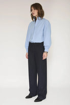 A model stands against a plain light background wearing a light blue long-sleeve button-up shirt with a high collar and dark navy pinstriped trousers.
