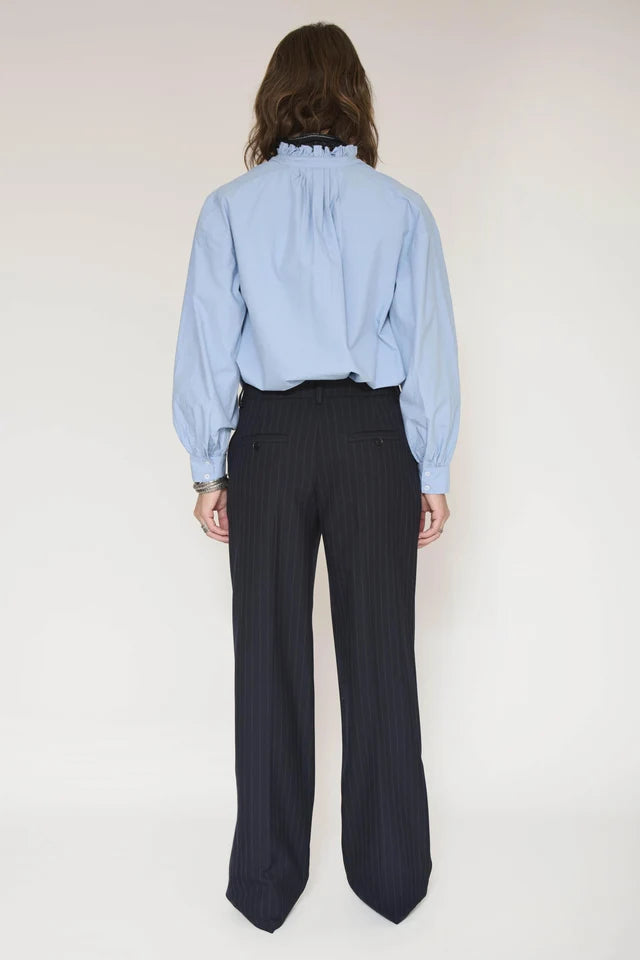 A model shown from the back wearing a light blue long-sleeve blouse with a ruffled high neck and dark navy pinstripe trousers, set against a plain off-white studio background.
