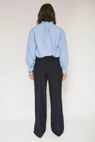 A model shown from the back wearing a light blue long-sleeve blouse with a ruffled high neck and dark navy pinstripe trousers, set against a plain off-white studio background.