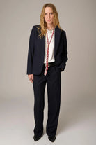A model stands against a neutral studio background wearing a dark navy pinstripe suit with a white shirt, a long red-and-white patterned scarf, and black shoes.