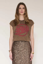 A woman modeling a brown cropped t-shirt with a red distressed 'The Strokes' logo, paired with a brown sequined skirt, against a plain light beige background.