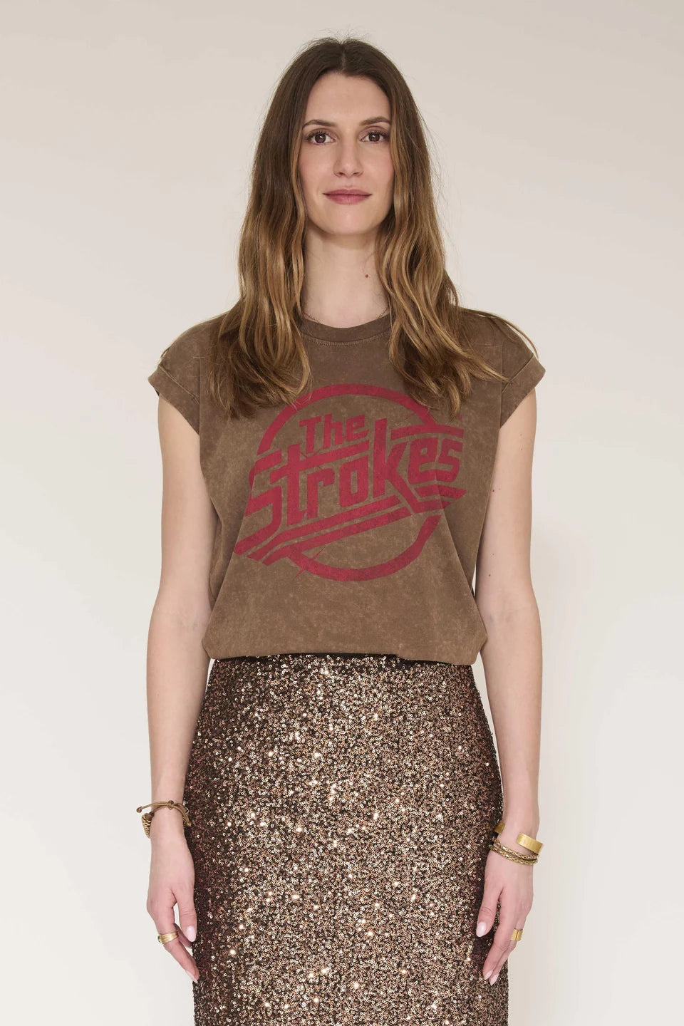 A woman modeling a brown cropped t-shirt with a red distressed 'The Strokes' logo, paired with a brown sequined skirt, against a plain light beige background.
