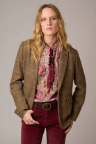 A woman modeling a brown plaid blazer over a pink and beige patterned blouse with a dark red necktie, paired with burgundy corduroy pants and a brown belt, against a solid gray studio background.