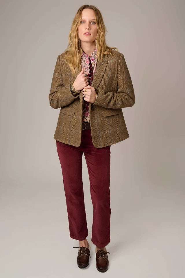 A woman modeling a brown plaid blazer, burgundy corduroy trousers, a patterned blouse, and dark brown leather shoes against a neutral gray studio background.