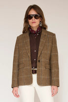 A woman modeling a brown and beige plaid tweed blazer over a dark brown button-up shirt, a burgundy patterned scarf, white pants, and sunglasses, against a plain light beige background.