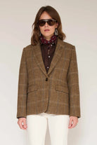 A woman wearing a brown plaid blazer over a dark brown button-up shirt with a burgundy patterned scarf, styled with white pants and dark sunglasses, against a plain light background.