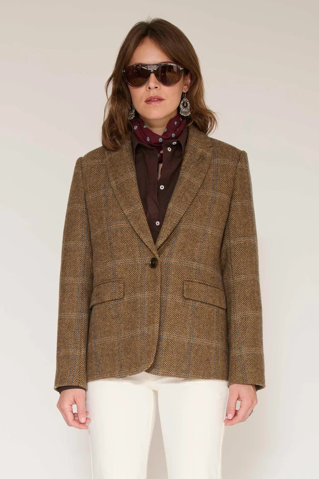 A woman wearing a brown plaid blazer over a dark brown button-up shirt with a burgundy patterned scarf, styled with white pants and dark sunglasses, against a plain light background.