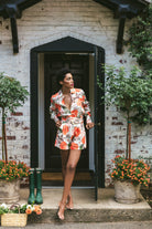 Woman in a floral dress standing on a porch with potted plants and a white brick wall.