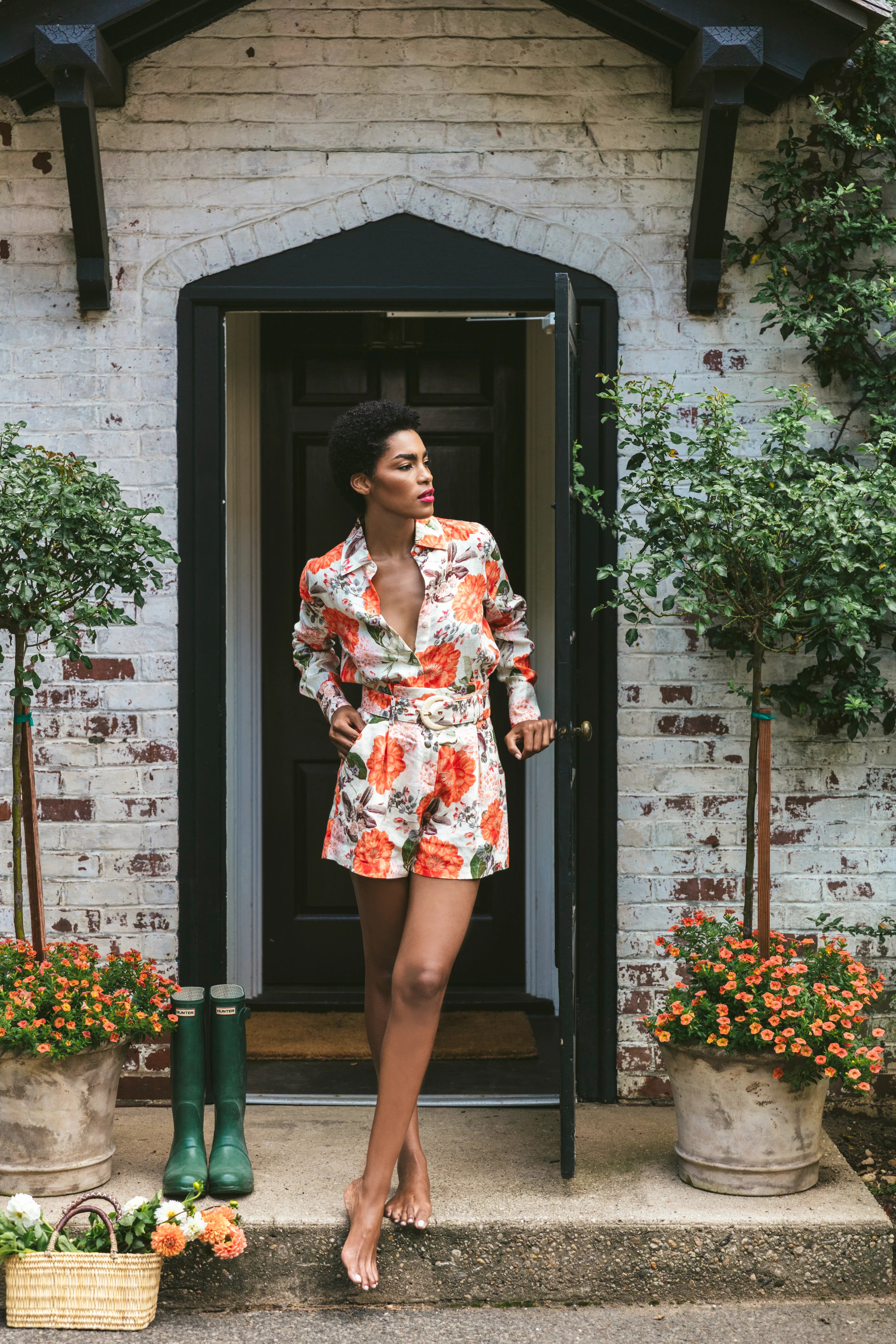 Woman in a floral dress standing on a porch with potted plants and a white brick wall.