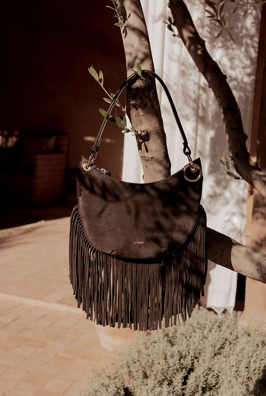 Brown leather handbag with fringe details hanging on a tree branch outdoors.