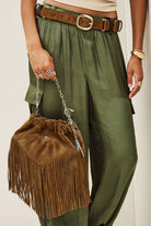 A person wearing olive green wide-leg pants and a brown belt, holding a brown suede fringe bucket bag with a silver chain strap.