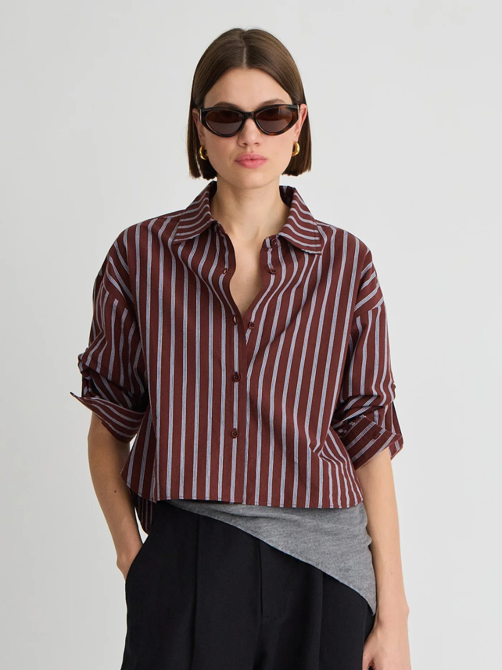 A model wearing a cropped, long-sleeve button-up shirt with vertical burgundy and gray stripes, styled with a gray underlayer, black trousers, and dark sunglasses against a light gray studio background.