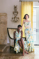Two women in floral dresses posing in a room with yellow curtains and decorative wall elements.