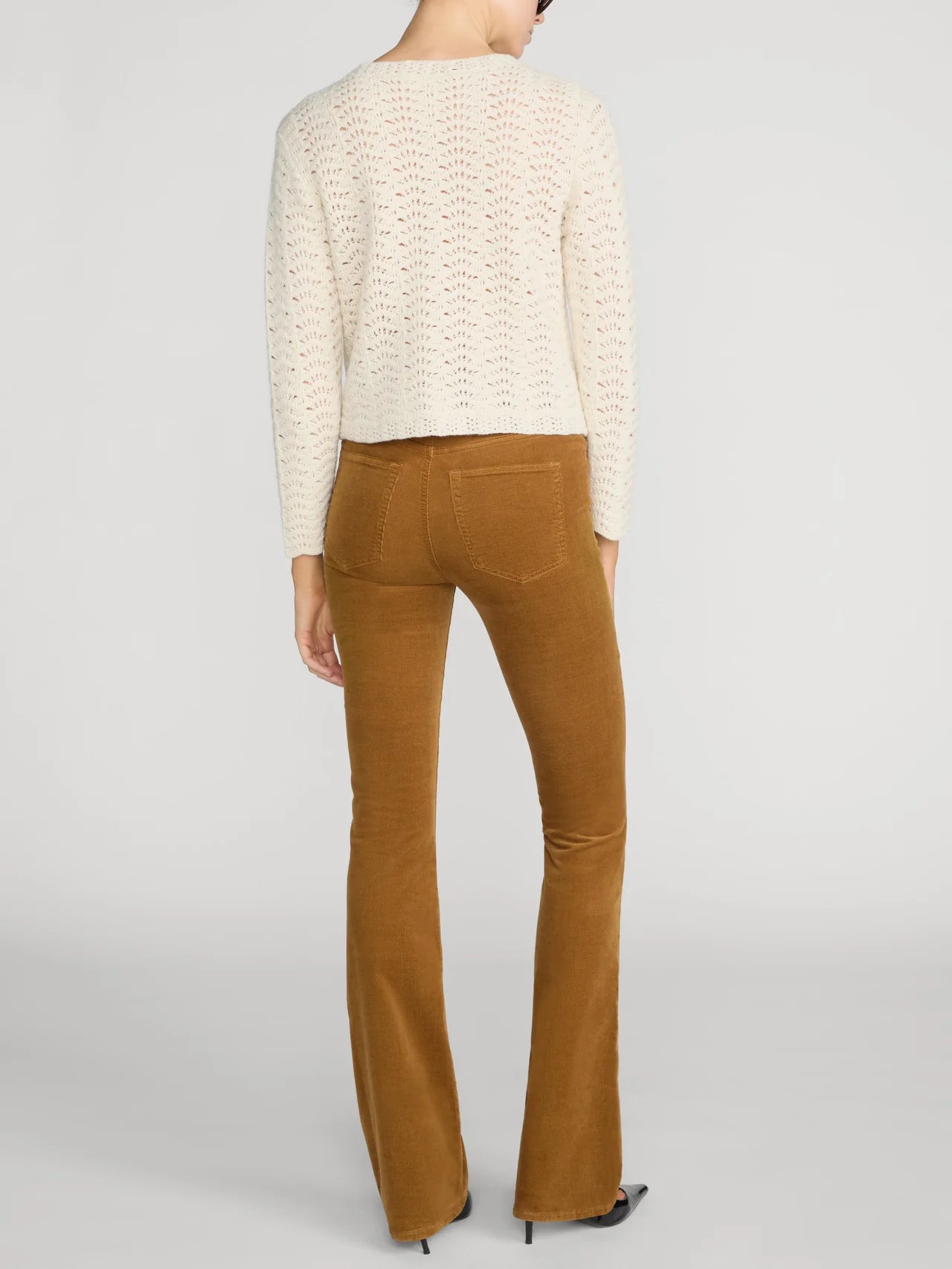 A model shown from the back wearing a cream-colored textured knit sweater and brown flared pants, set against a plain light gray studio background.