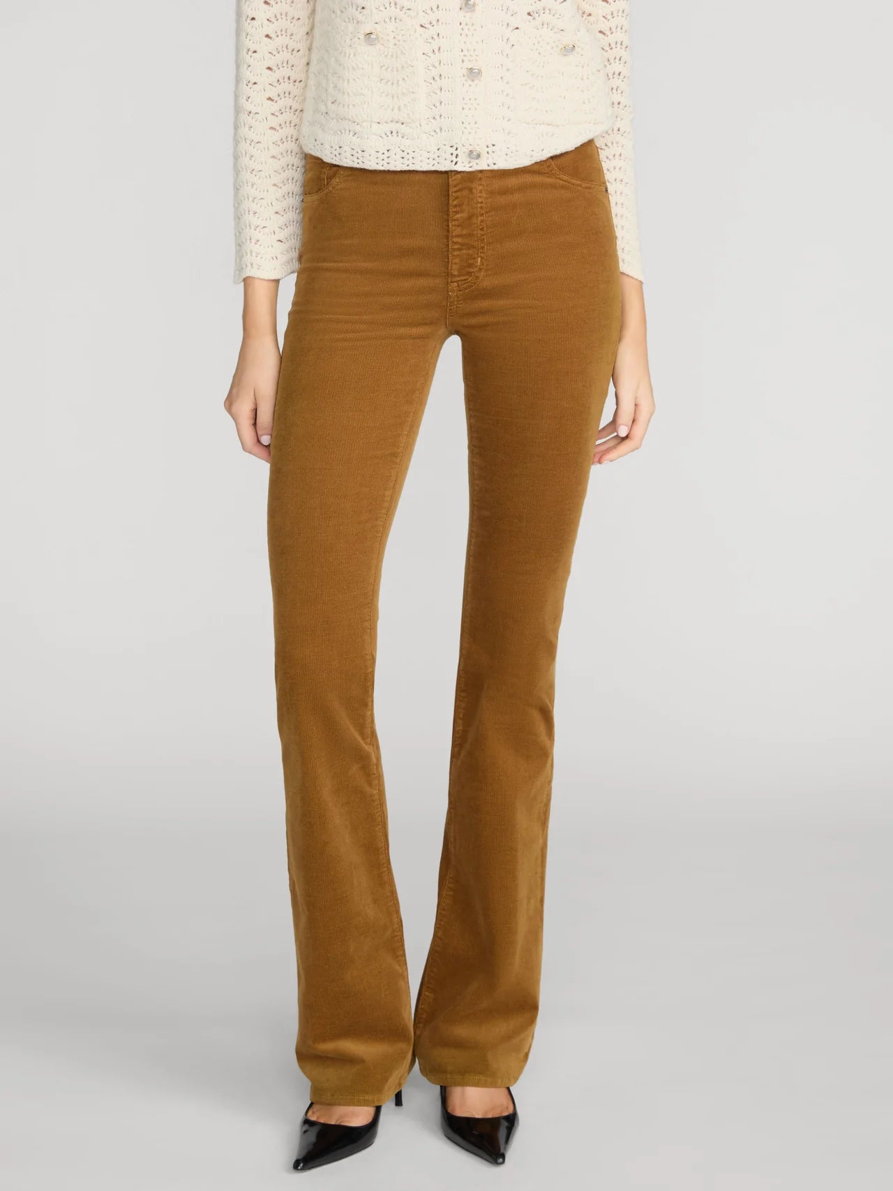 A model wearing a pair of brown corduroy flare pants, styled with a cream knit sweater and black pointed-toe heels, against a light gray studio background.
