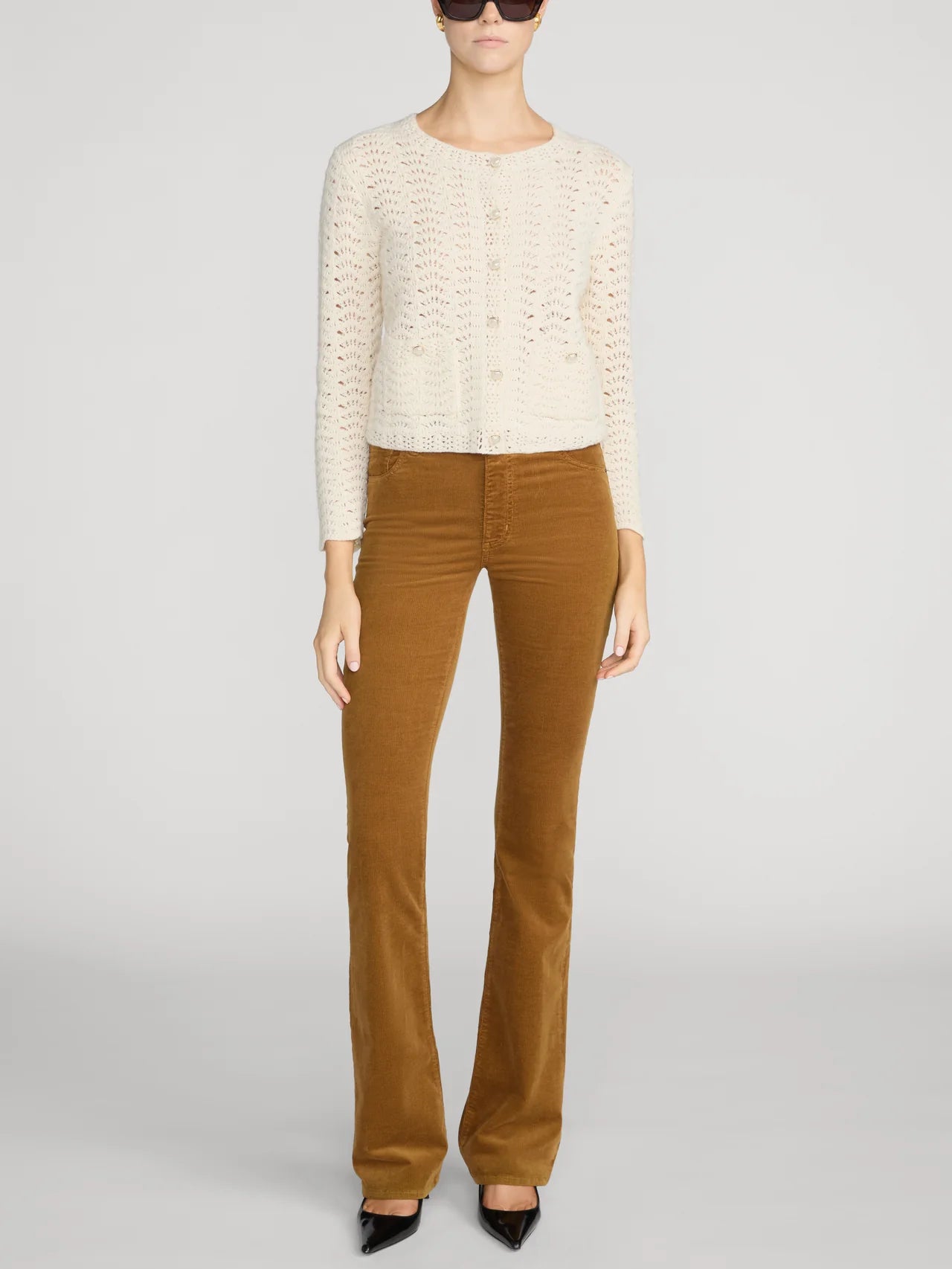 A model wearing a cream-colored textured knit cardigan, brown corduroy flare pants, and black pointed-toe heels, photographed against a neutral light gray studio background.