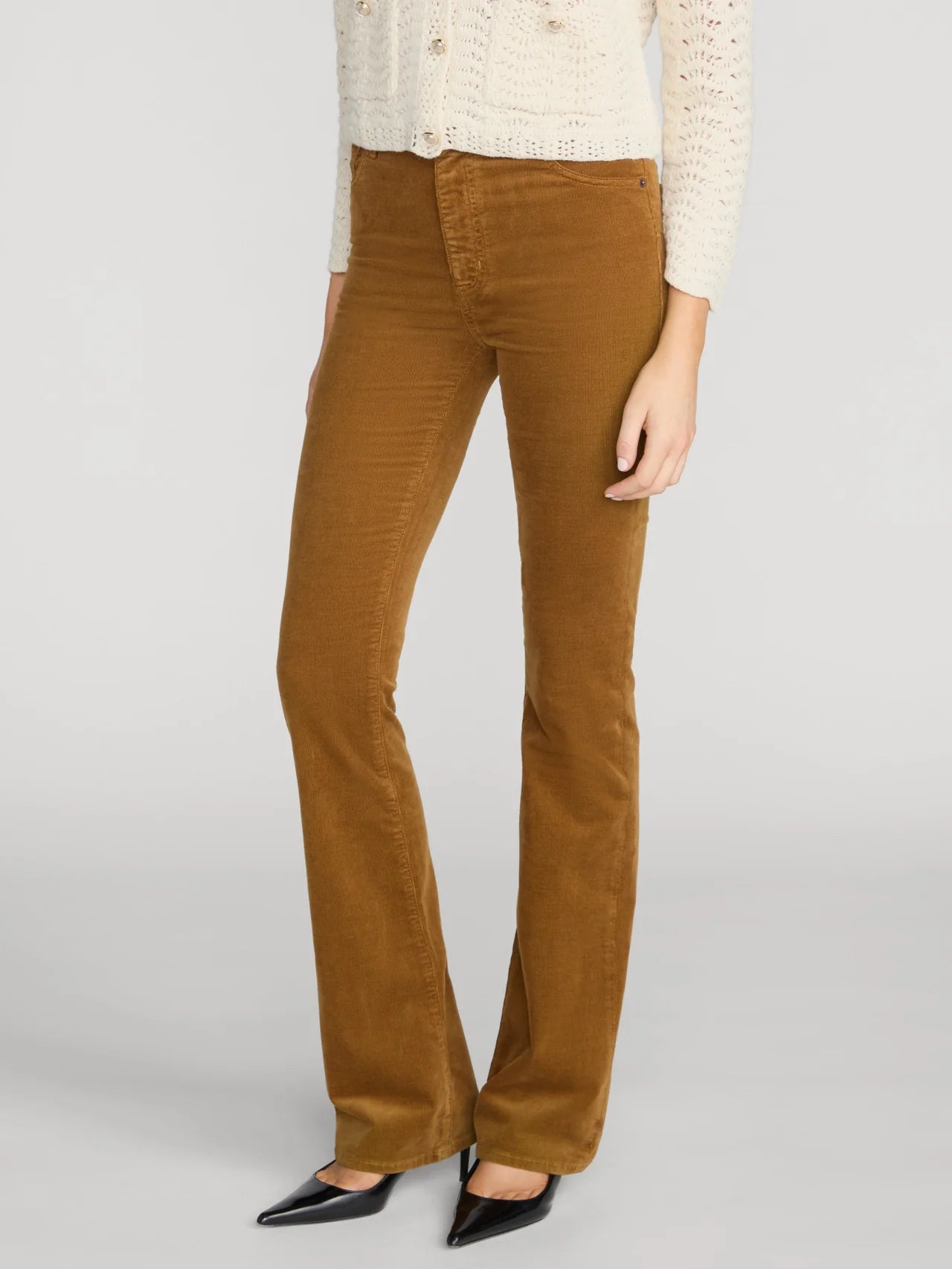 A model wearing a pair of brown corduroy flare pants, styled with a cream knit cardigan and black pointed-toe flats, against a light gray studio background.
