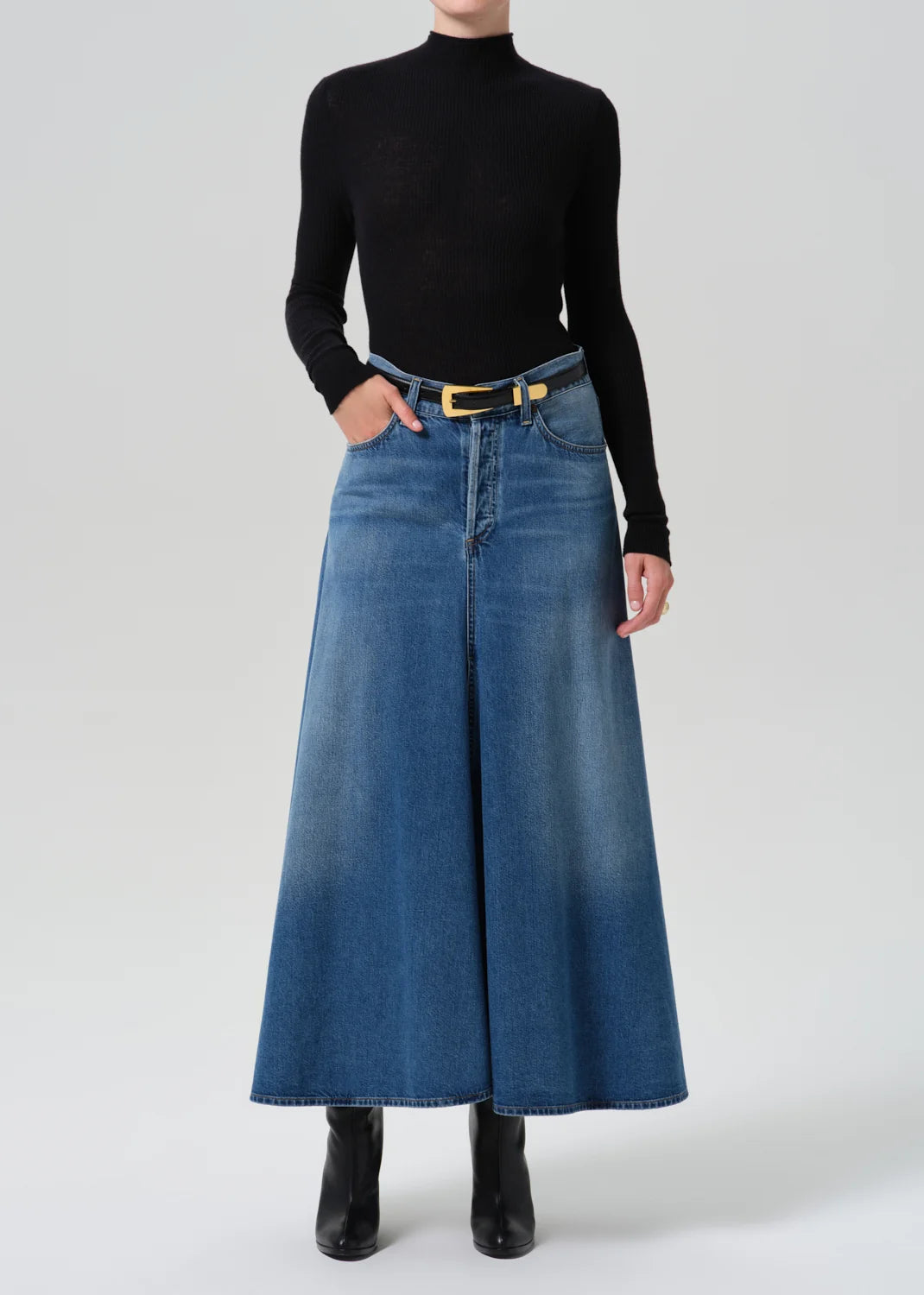 A model wearing a black long-sleeve mock neck top and a high-waisted, wide-leg blue denim skirt with a yellow-gold belt, paired with black ankle boots, against a light gray studio background.