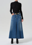 A model wearing a black long-sleeve mock neck top and a high-waisted, wide-leg blue denim skirt with a yellow-gold belt, paired with black ankle boots, against a light gray studio background.