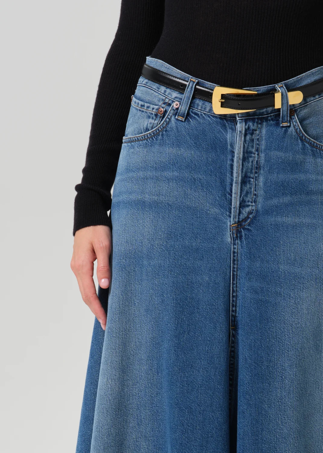 Close-up of a person wearing a blue wide-leg denim skirt with a black belt featuring a gold rectangular buckle, paired with a black long-sleeve top, against a light gray studio background.