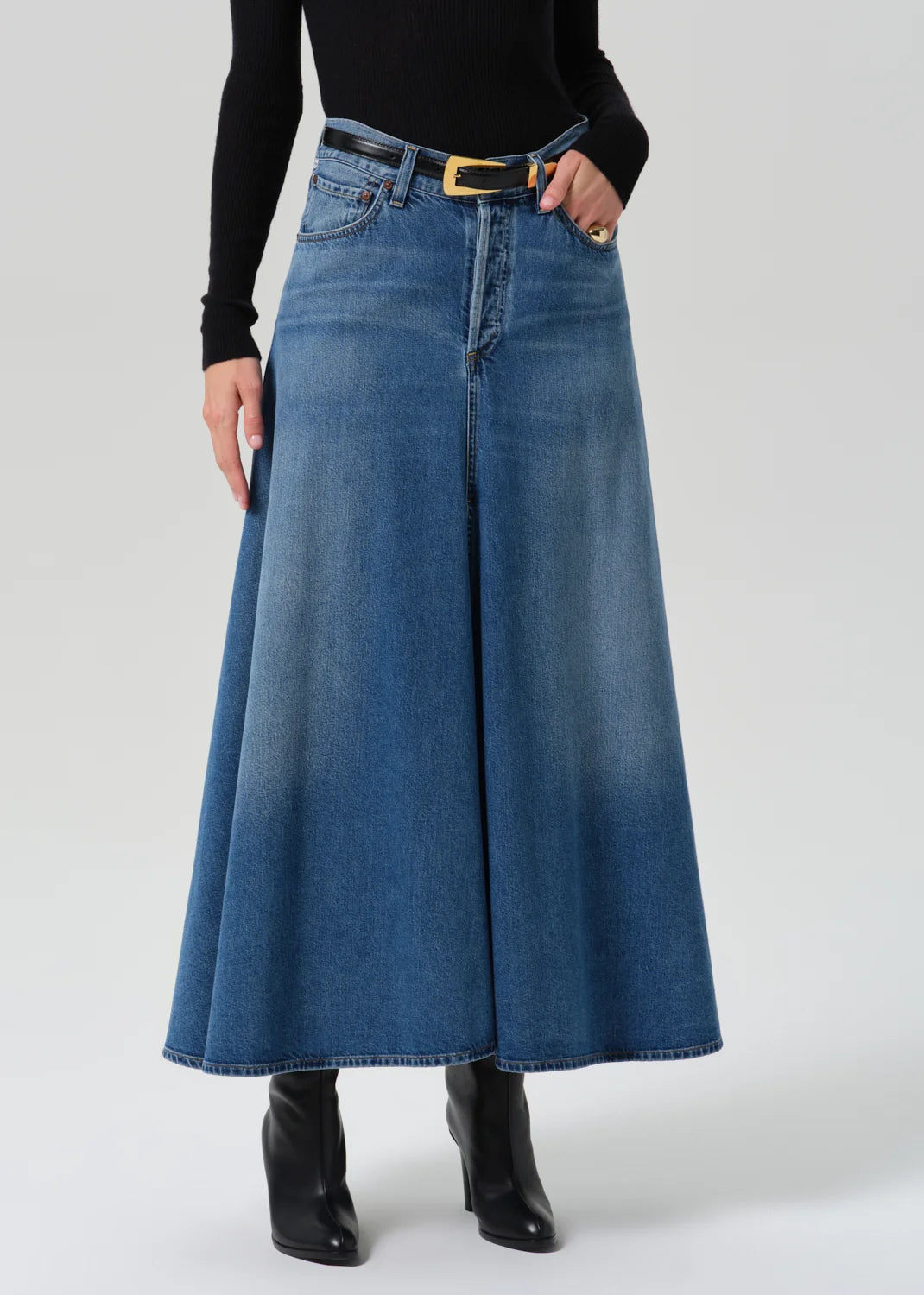 A model wearing a high-waisted, wide-leg blue denim skirt with a black belt and gold buckle, styled with a black long-sleeve top and black ankle boots against a light gray studio background.