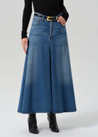 A model wearing a high-waisted, wide-leg blue denim skirt with a black belt and gold buckle, styled with a black long-sleeve top and black ankle boots against a light gray studio background.